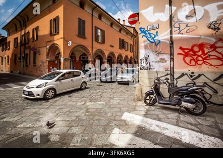 Bologna, Italien - 17. Mai 2018: Blick auf eine Kopfsteinpflasterstraße in Bologna mit historischen Säulenganggebäuden, geparkten Autos, einem Roller und Graffiti auf der C Stockfoto