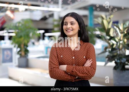 Junge professionelle Frau lächelt selbstbewusst mit verschränkten Armen, schaut weg in einem hellen modernen Firmenbüro, bereit und erfolgreich in professioneller Kleidung, vielfältig und entschlossen Stockfoto