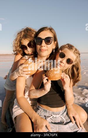 Zufriedene Mutter und zwei Töchter essen frische Sesamburger zusammen während eines sonnigen Familienpicknicks am Ufer des Salzsees und genießen köstliches Essen Stockfoto
