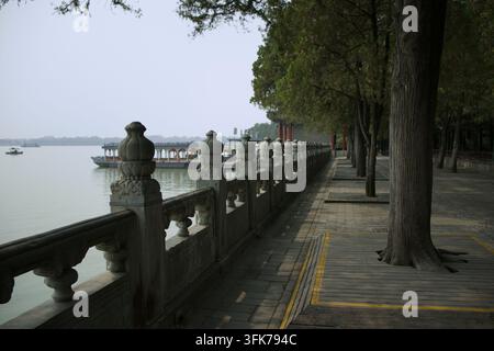 Langes SteinGeländer mit geschnitzten Balustern entlang eines Holzsteigs am Kunming-See, mit Weidenbäumen und Vergnügungsbooten, Sommerpalast, Peking, China Stockfoto