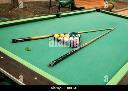 Billardtisch mit abgenutzter, fleckiger grüner Tischdecke, farbige Poolbälle im Dreiecksrack, Queue-Stöcke im Parkbereich Stockfoto