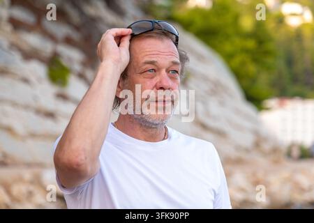 Ein Mann mittleren Alters in einem weißen Hemd hebt seine Sonnenbrille im Freien und schaut weg. Stockfoto