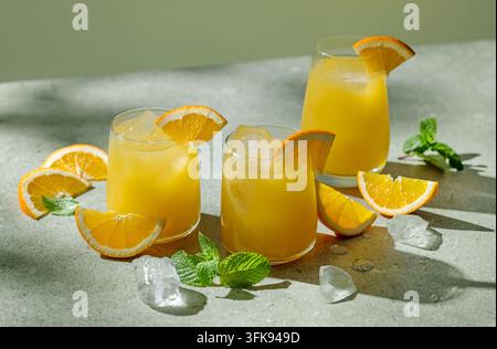 Drei Gläser gefüllt mit Orangensaft und Eiswürfeln, die auf einem sonnigen grauen Tisch mit frischen Orangenscheiben arrangiert sind. Erfrischendes Zitrusgetränke-Konzept su Stockfoto