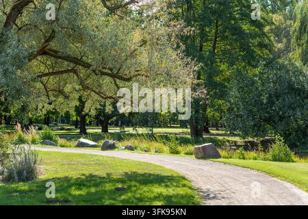 Ein sonniger gepflasterter Weg windet sich durch einen gestalteten Park mit ausgewachsenen Bäumen und grünem Gras. Stockfoto