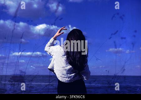 Künstlerisches blau getöntes Porträt einer Frau am Meer, von hinten betrachtet, mit botanischen Silhouetten, Vögeln und weichen Wolken, die eine surreale Atmosphäre schaffen Stockfoto
