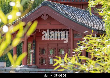 Roter traditioneller chinesischer Pavillon mit verzierten Holztüren und Ziegeldach, gesehen durch sonnenbeschienenes grünes Laub in einem friedlichen Garten. Stockfoto