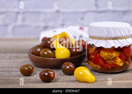 Glasbehälter mit getrockneten roten, schwarzen und gelben Kirschtomaten in Olivenöl und mit Kräutern auf dem rustikalen braunen Holztisch. Verschiedene Kirschtomaten in einer Stockfoto