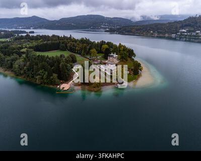 Luftbild der Maria-Worth-Halbinsel am Wörthersee mit historischer Architektur, üppigen Bäumen und ruhigem türkisfarbenem Wasser unter einer bewölkten Himmel Stockfoto