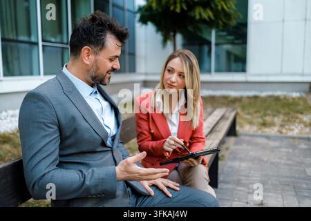 Zwei Fachleute sitzen auf einer Bank draußen in einem Geschäftsbereich und diskutieren, während sie ein Tablet nutzen. Stockfoto