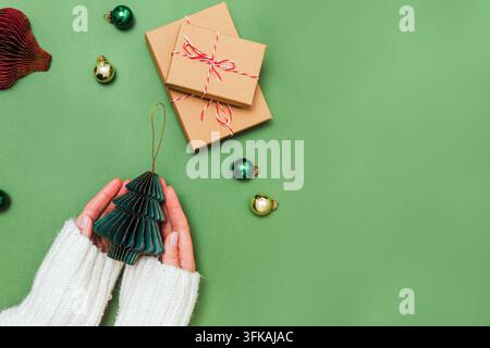 Weibliche Hände in einem kuscheligen weißen Strickpullover halten eine honigwabenförmige Weihnachtsbaumdekoration aus Papier neben Kraft-Geschenkboxen und Kugeln auf Grün. Stockfoto
