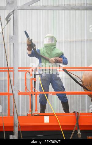 Ein Mann in Schutzkleidung bedient einen Sandstrahler von einer Scherenbühne aus, um Stahlarbeiten an einem feuerbeschädigten Gebäude zu reinigen, bereit für die Neubemalung. Stockfoto
