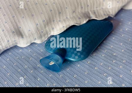 Eine blaue Warmwasserflasche, die auf einem Bett liegt und Wärme, Komfort und Entspannung bei kaltem Wetter darstellt. Warmwasserflaschen werden üblicherweise verwendet, um Stockfoto