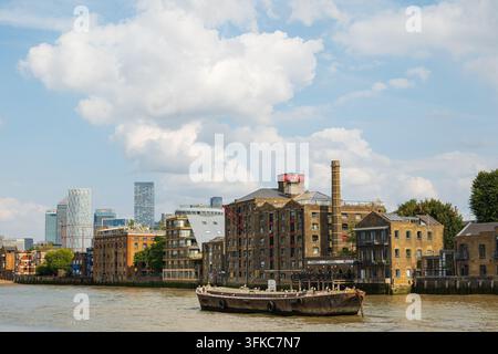 London, England, Vereinigtes Königreich, 16. August 2026. Historisches Thames Tunnel Mills-Gebäude und vertäutes Lastkahn auf der Themse mit Wolkenkratzern von Canary Wharf Stockfoto