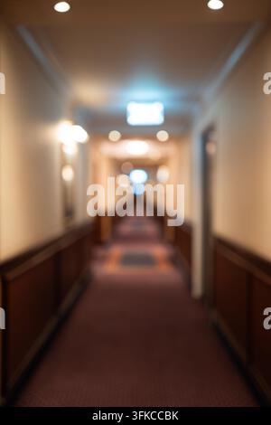 Unscharfer Innenblick auf einen langen Hotelkorridor mit Teppichboden, Wandvertäfelung und warmer Bokeh-Deckenbeleuchtung. Stockfoto