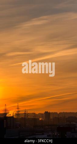 Vertikale Außenaufnahme bei Tageslicht eines dramatischen goldenen Sonnenuntergangshimmels mit gestreiften Wolken über einer dunklen urbanen Landschaft. Silhouette von Stadtgebäuden und Stromleitungen Stockfoto