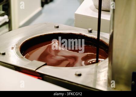 Geschmolzene Schokolade fließt aus einer Maschine in einer Küche. Dies ist ein üblicher Prozess bei der Dessertzubereitung, der Leckereien und Konfekte erzeugt. Stockfoto
