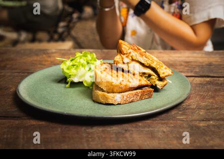 Frau, die ein gegrilltes Hühner-Panini-Sandwich mit einem Glas gekühltem Americano auf einem Holztisch genießt Stockfoto