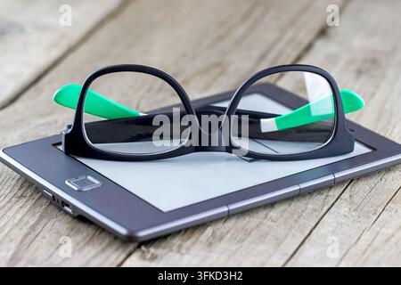Schwarze Brille, die auf einem Tablet auf einer Holzoberfläche ruht; die Komposition ruft Assoziationen mit Lesen, Technologie, Augenpflege und einem Arbeitsbereich hervor. Stockfoto