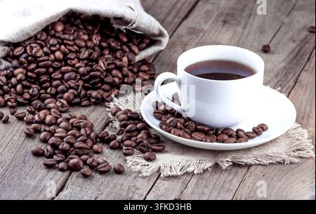 Frischer schwarzer Kaffee in einer weißen Tasse auf einer Untertasse neben Kaffeebohnen, die aus einem Jutesack auf einen Holztisch gefallen sind; die Atmosphäre eines warmen, gemütlichen Cafés Stockfoto