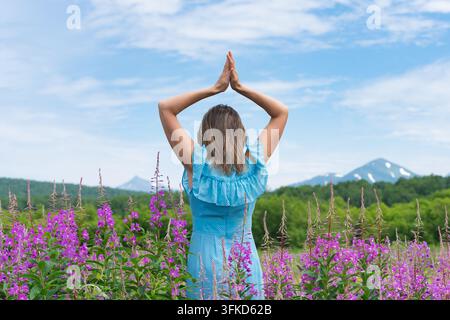 Frau in blauem Kleid steht mit erhobenen Händen über dem Kopf zwischen lila Wildblumen, Bergen und blauem Himmel im Hintergrund. Sommerliche Außenszene mit Ruhe Stockfoto