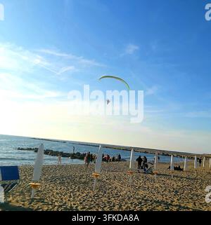 Eine Weitwinkelaufnahme eines Paramotors, der am hellblauen Himmel über dem Strand von Castel Volturno fliegt, während Menschen den sonnigen Küsten-Nachmittag genießen. Stockfoto