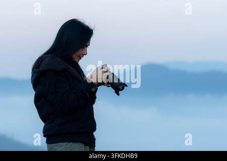 Eine junge Frau steht auf einem Berggipfel und fotografiert die malerische Landschaft während eines hellen Sonnenaufgangs am Morgen. Stockfoto