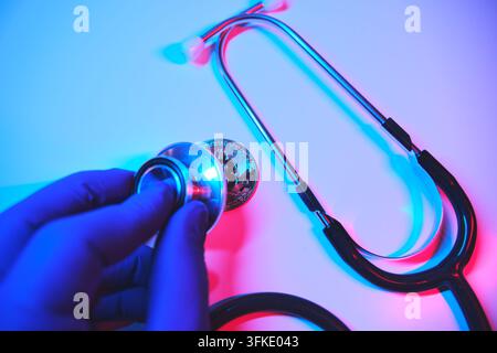 Hand in medizinischem Handschuh, die ein Stethoskop zu Bitcoin hält, symbolisch für Marktgesundheitsanalyse Stockfoto