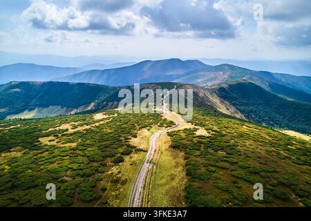 Luftaufnahme eines sich windenden Feldweges, der sich durch eine lebendige Berglandschaft schlängelt, mit üppigen grünen Sträuchern und Grasflächen, die zu fernen blau getönten Gipfeln unter einer teilweise bewölkten Himmel führen. Stockfoto