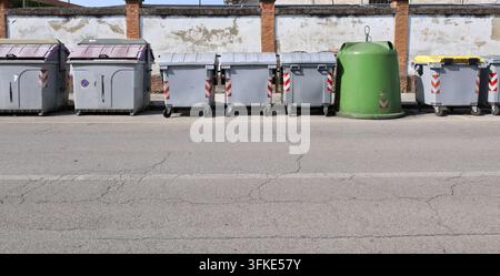 Gruppe von Mülltonnenbehältern in verschiedenen Größen am Straßenrand. Verwitterte Grenzmauer dahinter, Straße davor. Hintergrund für Kopierraum. Stockfoto