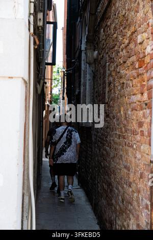 Zwei Personen gehen durch eine sehr schmale Gasse in Venedig zwischen verwitterten Ziegelmauern und weiß verputzten Wänden davon. Stockfoto