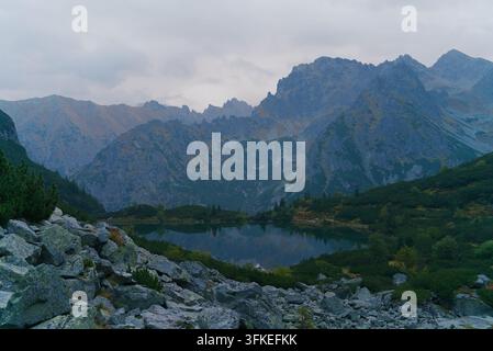 Ein ruhiger Alpensee spiegelt majestätische Berggipfel in einer friedlichen, felsigen Landschaft mit immergrüner Vegetation unter einem bewölkten Himmel wider. Stockfoto