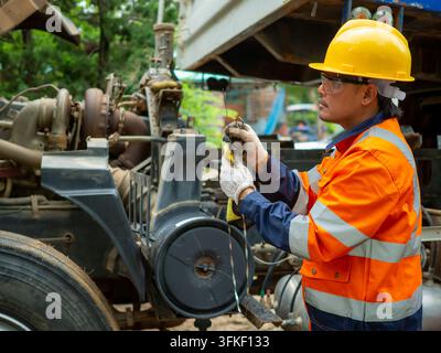 Ein asiatischer Mechaniker trägt einen gelben Helm und Sicherheitshandschuhe und prüft den Motorölstand mit einem Ölmaßstab während der Wartung eines schweren Lkw im Freien Stockfoto