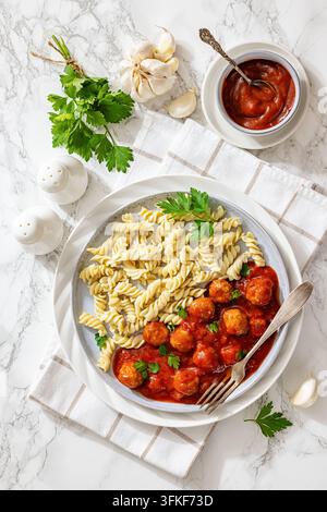 Klassisches amerikanisches Nudeln-und-Fleischbällchen-Gericht mit roter Tomatensoße, gedrehten Rotini-Nudeln, frischen Kräutern und Soßenschüssel auf einem dekorierten Tischtuch b Stockfoto