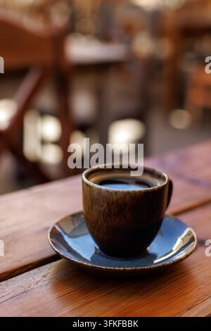 Eine braune Keramiktasse mit heißem schwarzem Kaffee auf einer Untertasse steht auf einer Holztablette in einem gemütlichen Außencafé-Setup. Stockfoto