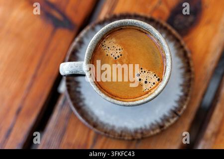 Eine keramische Tasse frisch gebrühten schwarzen Kaffees mit reichem Schaum steht auf einer Untertasse über einem warmtönigen Holztisch. Stockfoto