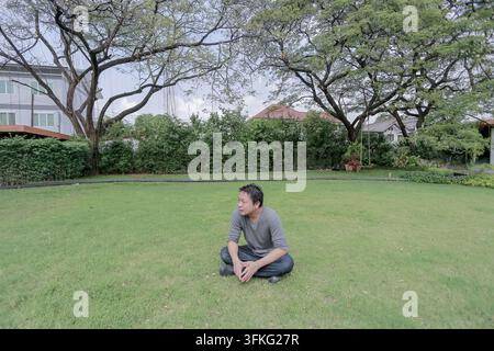 Asiatischer Mann sitzt im Schneidersitz auf einem grünen Rasen unter großen Bäumen. Er schaut weg, während er sich in einem ruhigen Garten entspannt. Stockfoto