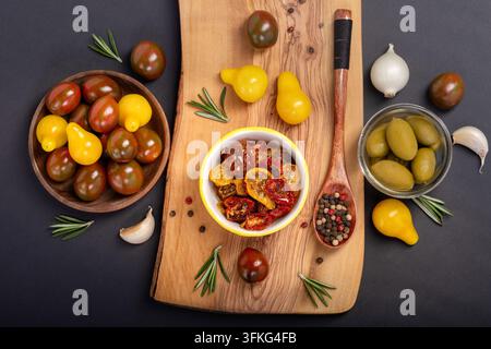 Stillleben-Foto eines Holzbretts auf schwarzem Hintergrund mit sonnengetrockneten und frischen Kirschtomaten, eingelegten Oliven in Untertassen, frischem Rosmarin und Pfefferkörnern Stockfoto
