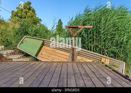 Hölzerner Steg und altes Boot am Ufer, überwuchert mit grünem Schilf am Wasser, friedliche Naturlandschaft, Landschaft. Stockfoto