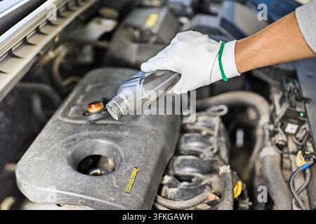 Nahaufnahme eines Mechanikers mit Handschuhen, der während der Wartung Motoröl in einen Automotor unter der Haube gießt. Stockfoto