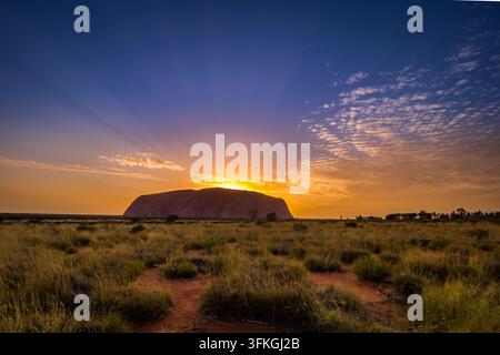 Uluru, Australien – 21. Januar 2019: Die aufgehende Sonne sendet Strahlen über Uluru (Ayers Rock) in einen farbenfrohen Himmel mit Wolken über einer Spinifex-Ebene Stockfoto