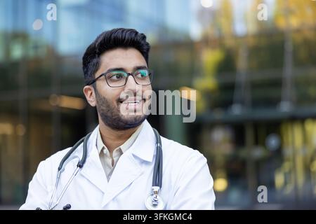 Junger indischer Arzt trägt einen weißen Laborkittel und Stethoskop, lächelt und schaut mit einem nachdenklichen Ausdruck weg, repräsentiert Gesundheitswesen und medizinische Innovation Stockfoto