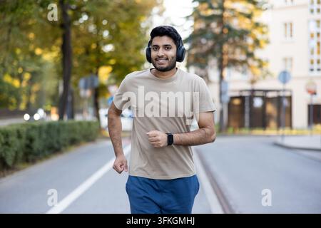 Junger Mann joggt in einem Stadtpark, lächelt mit Kopfhörern und Smartwatch, genießt einen sonnigen Tageslauf, gesunder aktiver Lebensstil und energiegeladener Outdoor-Workout Stockfoto