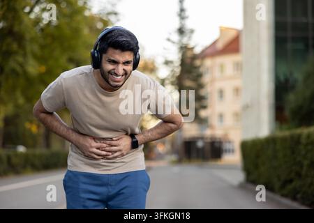 Junger Mann mit Kopfhörern und Sportkleidung, der sich mit beiden Händen den Bauch hält und unter starken Bauchschmerzen oder einem Seitenstechen während eines Trainings in einer Parkumgebung leidet Stockfoto