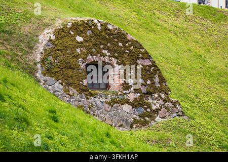 Ein mehrteiliges Fenster, umrahmt von verwittertem Ziegelmauerwerk und moosbewachsenen Steinen, ist in die Seite eines steilen, grasbewachsenen Hügels eingebaut. Das umgebende Mauerwerk i Stockfoto
