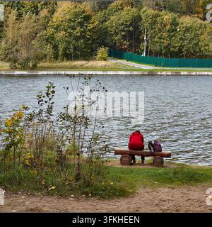 Person, die eine rote Jacke trägt, sitzt auf einer rustikalen Bank neben plätscherndem Wasser, mit einer Tasche in der Nähe, Herbstvegetation, Bäumen und einem fernen Zaun Stockfoto