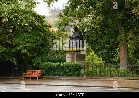 Edinburgh, Schottland - 3. August 2026: Regenreicher Park mit sitzender Statue auf einer Bank neben Bäumen und einem Zaun, mit entferntem Schloss auf einem Hügel unter wolkigem Tageslicht. Stockfoto