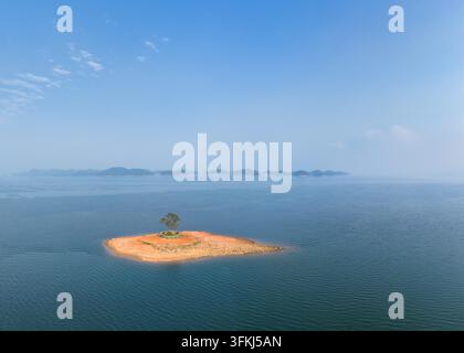 Luftaufnahme einer winzigen Insel mit einem einzigen Baum in der Mitte eines weiten, ruhigen Sees, umgeben von fernen Hügeln unter einem klaren blauen Himmel Stockfoto
