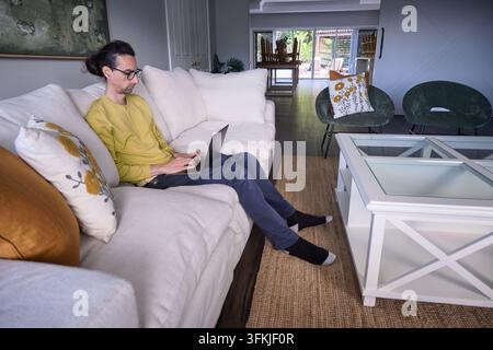 Mann arbeitet an einem Laptop in einem geräumigen Wohnzimmer mit Gartenblick Stockfoto