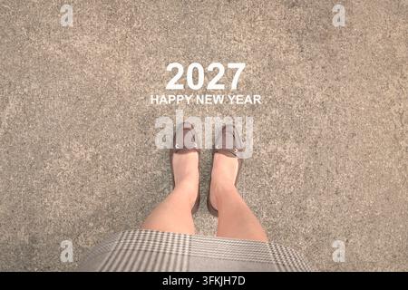2027 Glückliches Neujahrsgruß auf Asphaltoberfläche vor einer Geschäftsfrau mit braunen Absätzen. Vogelperspektive. Stockfoto