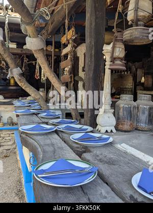 Pirovac, Kroatien, 16. August 2026: Ein verwitterter Holztisch ist für ein lockeres Abendessen mit weißen Tellern, blauen Servietten und silbernem Besteck gedeckt, umgeben von Stockfoto